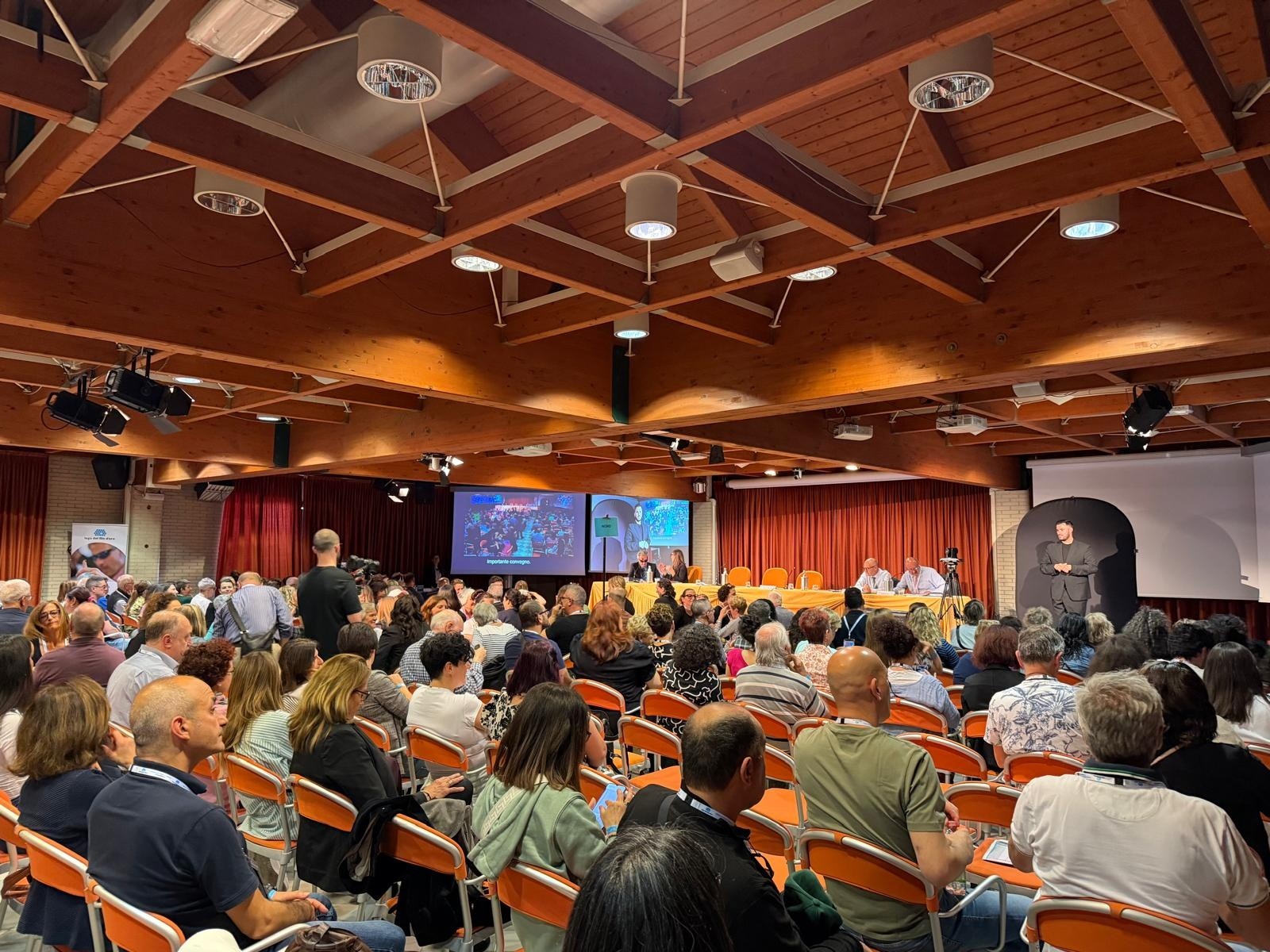 Foto dei partecipanti della X Conferenza delle Persone Sordocieche, tenutasi a Cervia dal 7 al 10 maggio 2026. I partecipanti sono riuniti in una ampia sala, disposti in più file, durante un momento dell’evento. Sullo sfondo il tavolo dei relatori, un interprete LIS sulla destra e due maxi schermi sulla sinistra. 