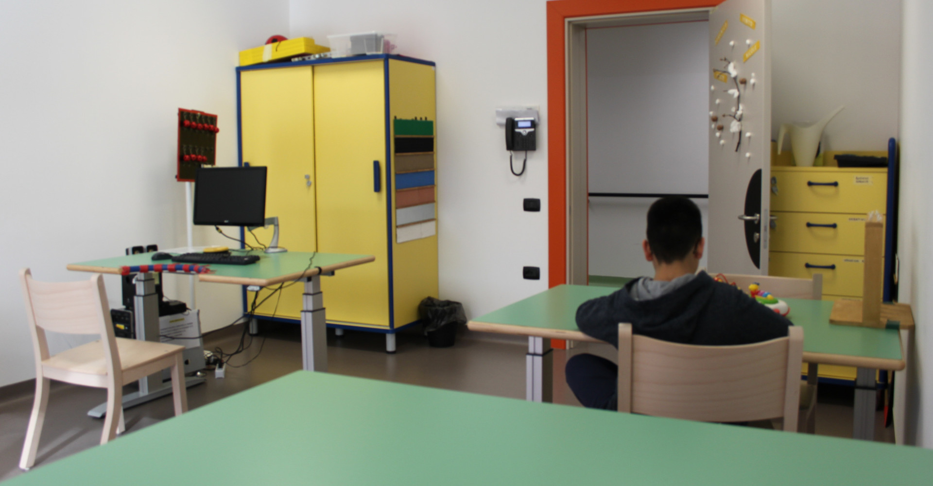Interno di un'aula per attività educativo-riabilitative del Centro Nazionale con diverse postazioni attrezzate. Si vede un computer su uno dei banchi, mentre un altro banco è occupato da un piccolo utente che sta svolgendo un'attività, visto di spalle