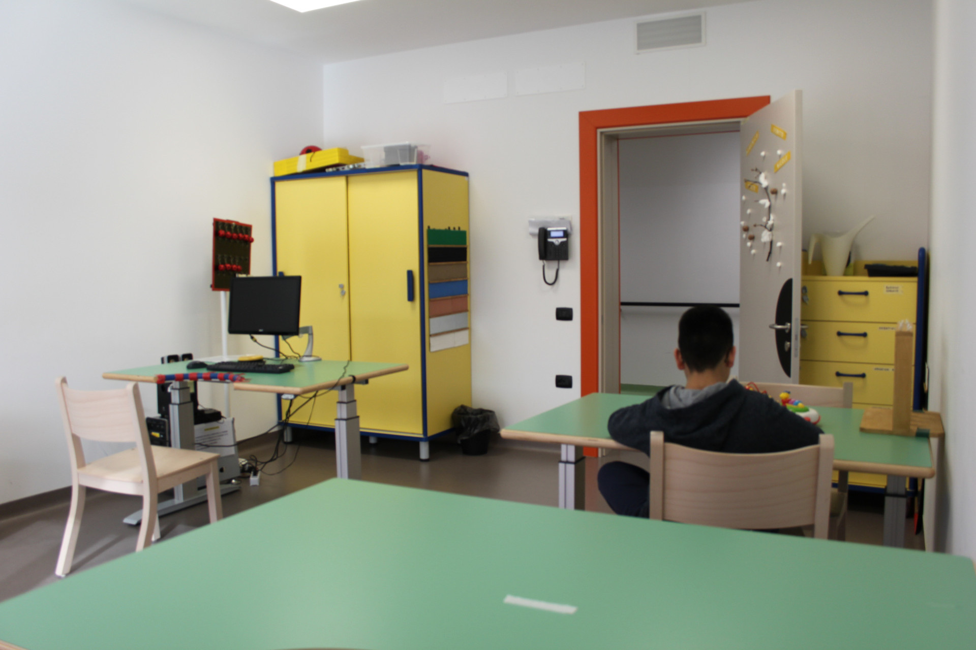 Interno di un'aula per attività educativo-riabilitative del Centro Nazionale con diverse postazioni attrezzate. Si vede un computer su uno dei banchi, mentre un altro banco è occupato da un piccolo utente che sta svolgendo un'attività, visto di spalle