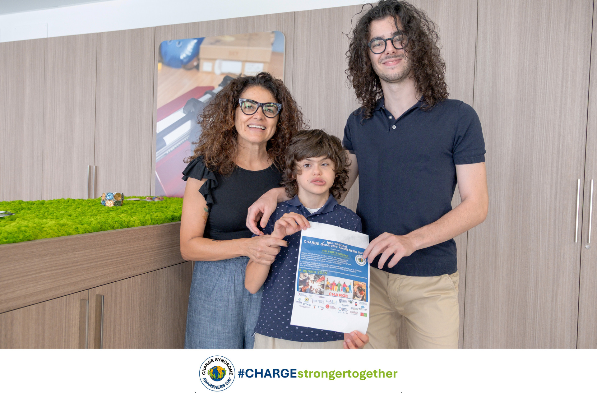 Agostino, ragazzo con la sindrome di CHARGE, posa insieme alla mamma e il fratello con la locadina del Charge Awareness Day che si festeggia l'8 Agosto.