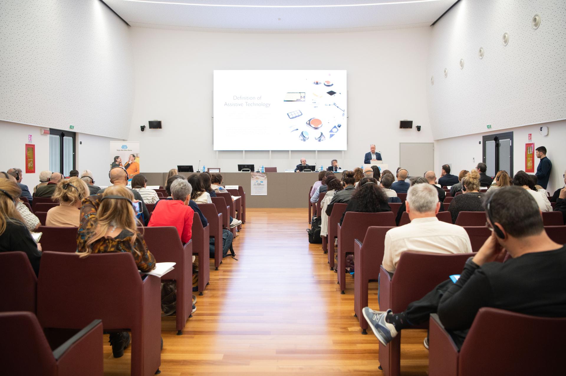 Terza Edizione della Conferenza Internazionale su Tecnologie Assistive e Disabilità. Auditorium della Lega del Filo d'Oro di Osimo gremito di persone sedute sulle poltrone che ascoltano la conferenza.