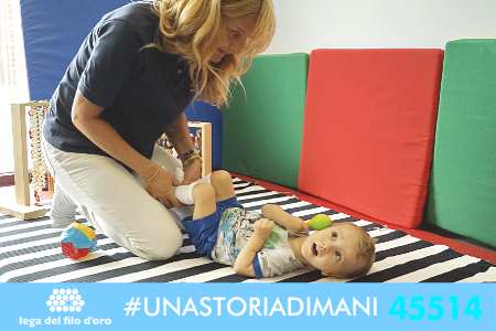 Immagine di campagna #UnaStoriaDiMani, con hashtag e numero di telefono in basso. Nell'immagine un'operatrice fa fisioterapia con un bambino.