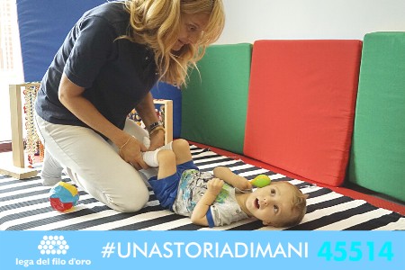 Immagine di campagna #UnaStoriaDiMani, con hashtag e numero di telefono in basso. Nell'immagine un'operatrice fa fisioterapia con un bambino.