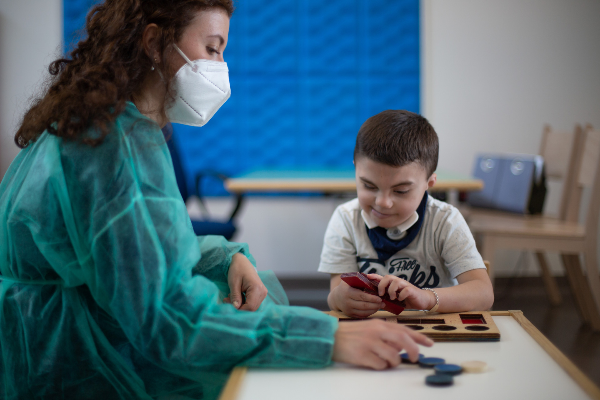 Leonardo sorride mentre ricompone con le sagome corrispondenti un puzzle di legno. Al suo fianco c'è la sua educatrice, che indossa camice monouso e mascherina ffp2