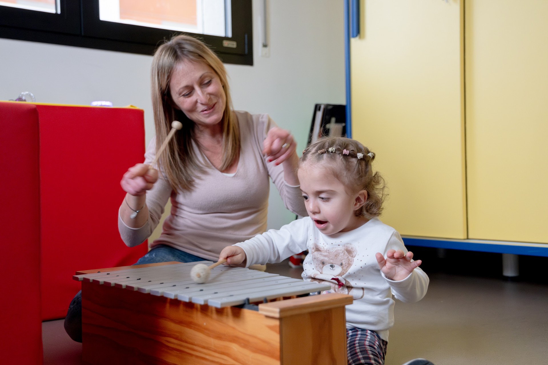 Una educatrice e una bambina della Lega del Filo d'Oro siedono a terra in un ambiente colorato e giocano con uno xilofono. Entrambe tengono in mano le bacchette e colpiscono le lamelle dello strumento. Sullo sfondo sono visibili le ante dei mobili e pannelli colorati.