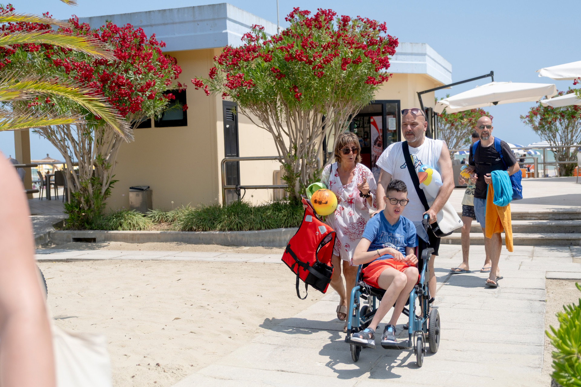 Un ragazzo in carrozzina, accompagnato da due volontari, rientra dalla spiaggia al soggiorno estivo di giugno 2025 a Giulianova: un momento di condivisione, cura e libertà.