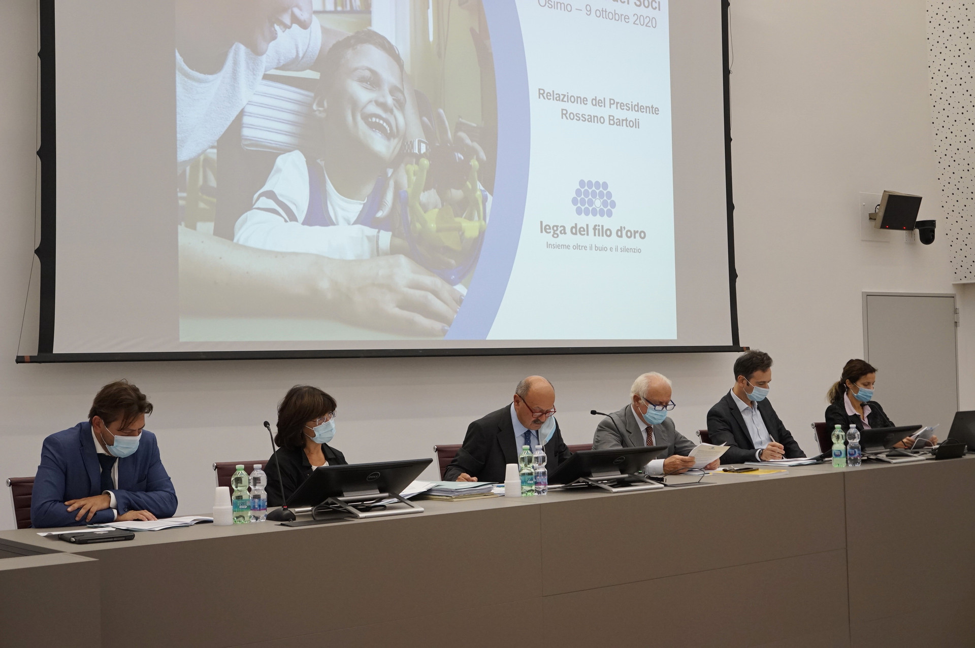 Cinque persone tra cui il Presidente Bartoli parlano ad una sala, mentre dietro di loro sono proiettate immagini di una presentazione.