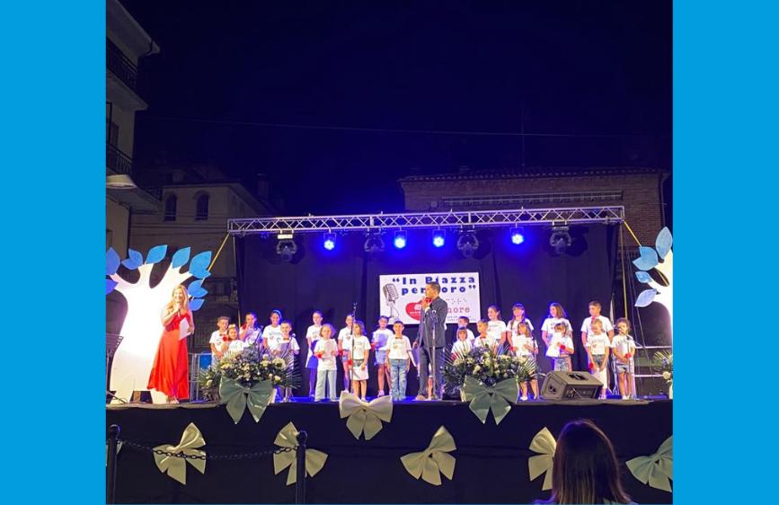 L'iimmagine mostra un palcoscenico allestito di notte, illuminato da luci blu, con una performance in corso. Sul palco, al centro, un gruppo di bambini e ragazzi in magliette bianche sta cantando, mentre ai lati ci sono due donne, una in abito rosso e l'altra che sembra presentare. Sullo sfondo, dietro i giovani, un pannello luminoso mostra la scritta "In Piazza per loro", e il palco è decorato con alberi stilizzati e grandi fiocchi bianchi, suggerendo un evento dedicato ai bambini o un'iniziativa di beneficenza.