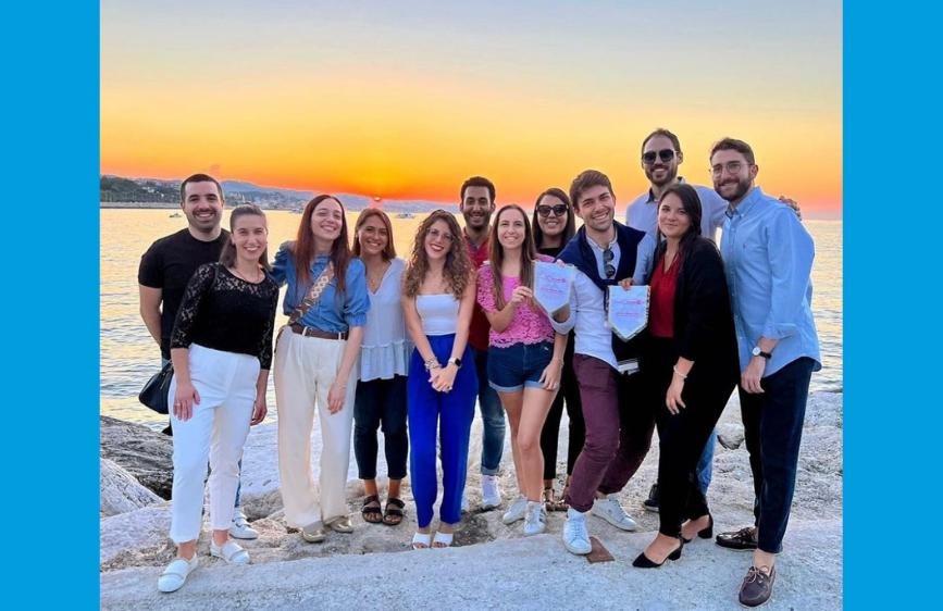 Questa foto mostra un gruppo di undici giovani adulti, membri del Rotaract, in posa su un molo o una scogliera rocciosa al tramonto. Tutti sorridono, alcuni indossano abiti semi-formali e due di loro reggono un gagliardetto con il simbolo del Rotaract
