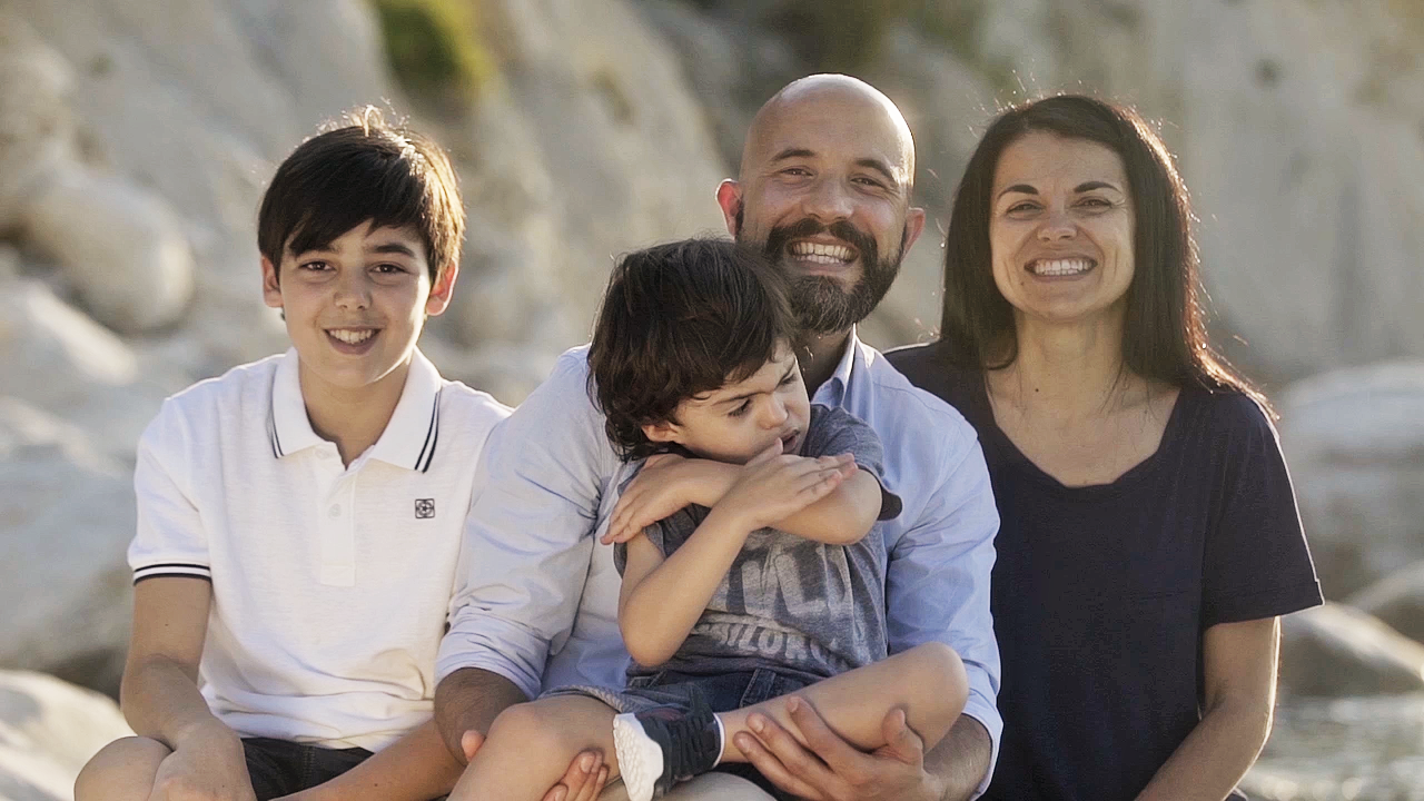 Foto della famiglia di Agostino, bambino con sindrome di Charge seguito a Osimo. Il papà lo tiene in braccio mentre il fratello maggiore e la mamma sorridono al suo fianco