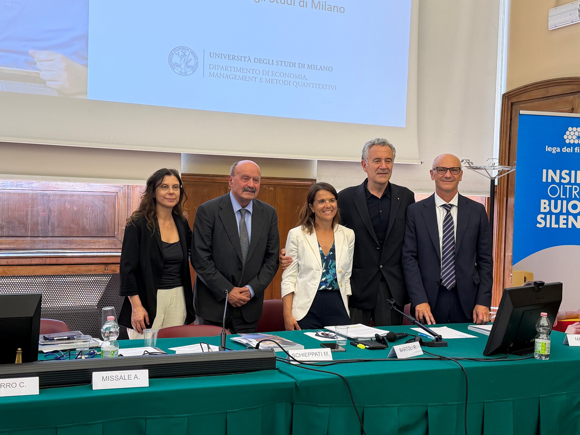 Da sinistra a destra: Barbara Duca, Direttore Amministrazione Finanza e Controllo Lega del Filo d’Oro, Rossano Bartoli, Presidente Lega del Filo d’Oro; Clodia Vurro, Responsabile scientifico del progetto, Università degli Studi di Milano; Enzo Manes, Fondatore Dynamo Camp; Roberto Costantini, Direttore Generale Lega del Filo d’Oro. Il gruppo è in posa dietro una scrivania, si trovano all'interno della Sala Lauree dell’Università degli Studi di Milano.