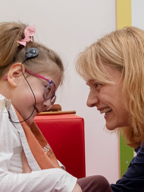 Eleonora, bambina sordocieca cospite del Centro Nazionale di Osimo, scambia un sorriso divertito con Francesca, la fisioterapista con cui sta svolgendo degli esercizi fisici. La bambina è seduta su dei grandi cuscini colorati e porta un impianto cocleare.