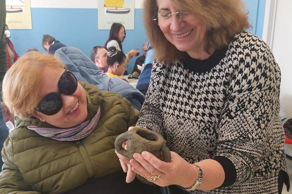 Paola, utente della Lega del Filo d'Oro, e una volontaria sono ritratte mentre modellano a mano una tazza di argilla.