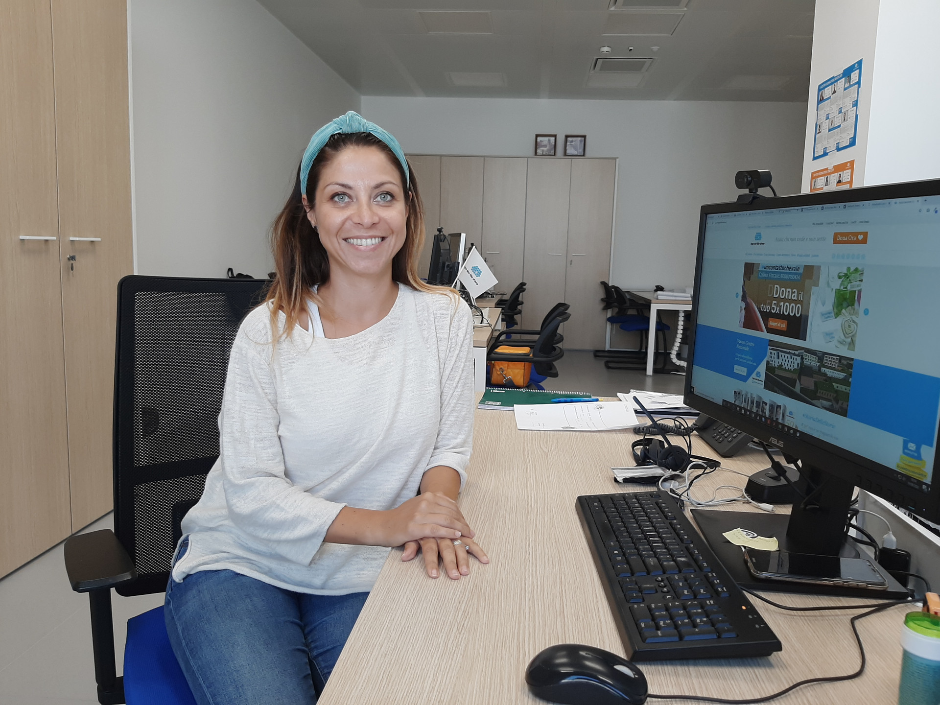 Rachele, responsabile Digital Marketing della Lega del Filo d'Oro, è seduta alla sua scrivania e sorride alla fotocamera. Sulla scrivania ci sono i materiali e gli strumenti per lavorare, come la cancelleria e il computer.