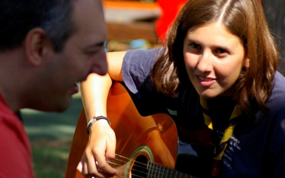 Una volontaria scout suona la chitarra per divertire un utente adulto del Centro di Osimo. Sono entrambi seduti all'aperto, nel giardino della sede storica della Lega del Filo d'Oro