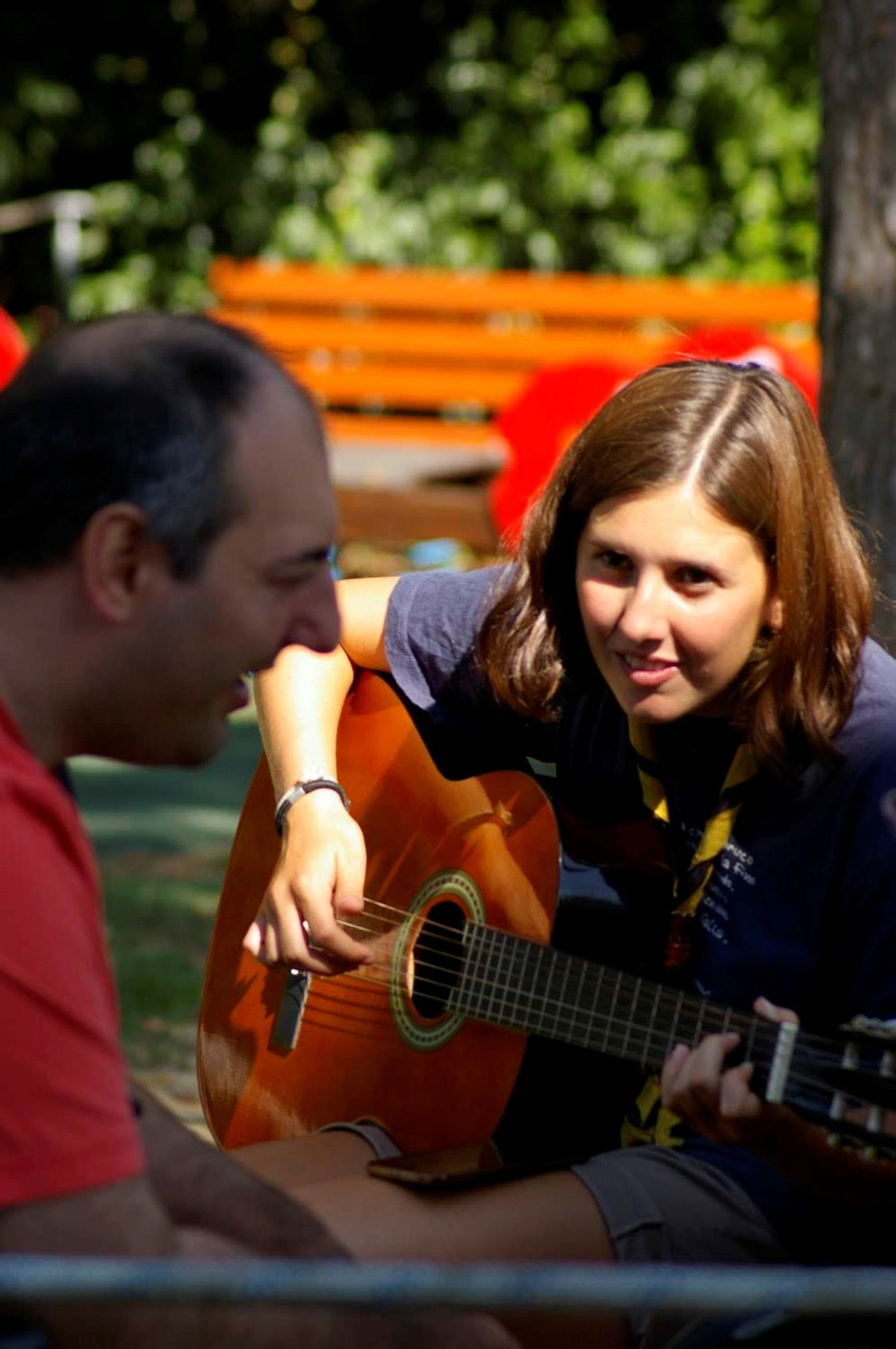 Una volontaria scout suona la chitarra per divertire un utente adulto del Centro di Osimo. Sono entrambi seduti all'aperto, nel giardino della sede storica della Lega del Filo d'Oro