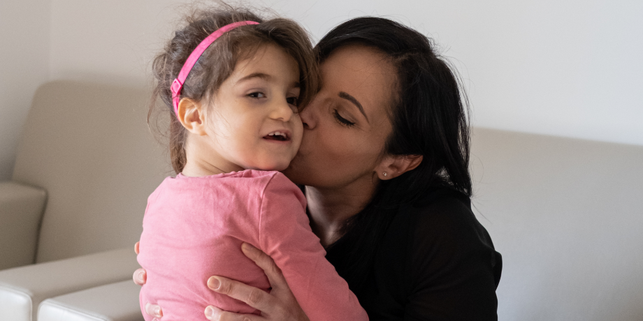 Mamma Alessandra con la piccola Letizia, bambina con la sindrome di Charge di circa 10 anni. La mamma la tiene sulle ginocchia, tenendola per la vita e le dà un bacio sulla guancia.