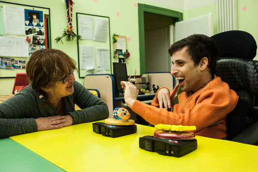 Un utente adulto sta comunicando con la sua educatrice grazie a un ausilio tecnologico. Lo strumento è composto da due pulsanti switch con riferimenti tattili, connessi a un sintetizzatore vocale.