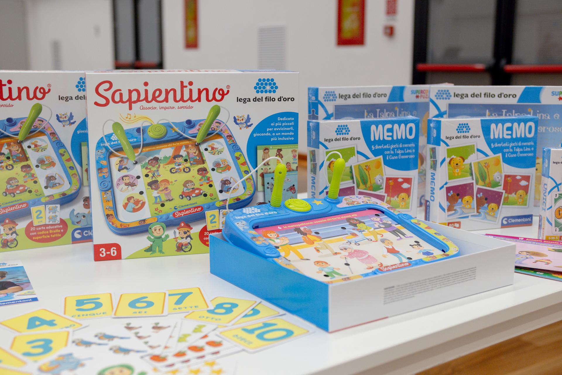 Su un tavolo compaiono 2 scatole chiuse dellla versione speciale del Sapientino, 2 scatole del gioco Memo e, delle carte numerate. Giochi realizzati da Clementoni per il catalogo di Natale della Lega del Filo d'Oro