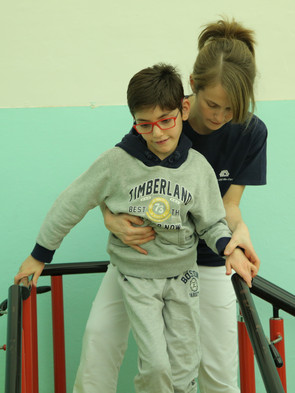 Federico, piccolo utente del centro di Osimo, durante una seduta di fisioterapia. Il ragazzino cammina tenendosi al corrimano lungo un percorso con salite e discese, mentre la fisioterapista lo assiste sorreggendolo.