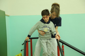 Federico, piccolo utente del centro di Osimo, durante una seduta di fisioterapia. Il ragazzino cammina tenendosi al corrimano lungo un percorso con salite e discese, mentre la fisioterapista lo assiste sorreggendolo.