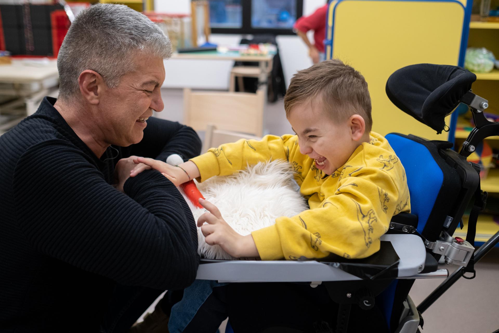 La foto ritare l'educatore Andrea mentre fa attività con Edoardo e ridono insieme. Andrea ha appoggiato sul tavolino della sedia di Edoardo un tappetto peloso per fargli sentire con il tatto la consistenza.