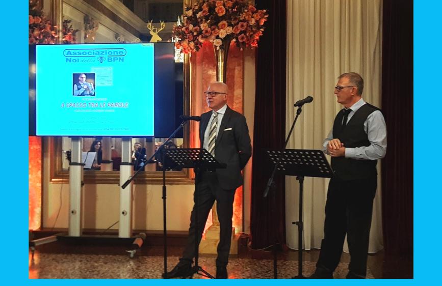 L'immagine mostra due uomini sul palco durante un evento formale, con un grande schermo retrostante che proietta il logo di "Associazione Noi BPN". L'uomo a sinistra, vestito in abito scuro e cravatta, sta parlando al microfono, mentre l'uomo a destra, in gilet, lo ascolta. Sullo sfondo si notano decorazioni floreali e tendaggi.