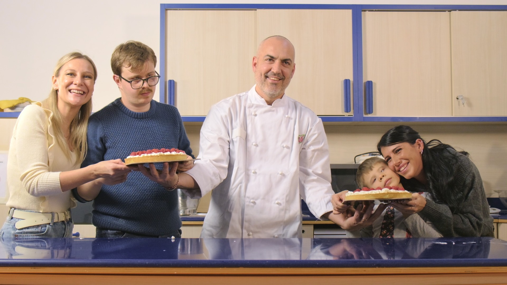 Un pasticcere nella cucina didattica (presso il Centro Nazionale della Lega del Filo d'Oro) con Victor e la sua operatrice, Matteo con la sua educatrice. Sono in posa per la foto di gruppo e le loro torte con la panna montata appena preparate insieme.