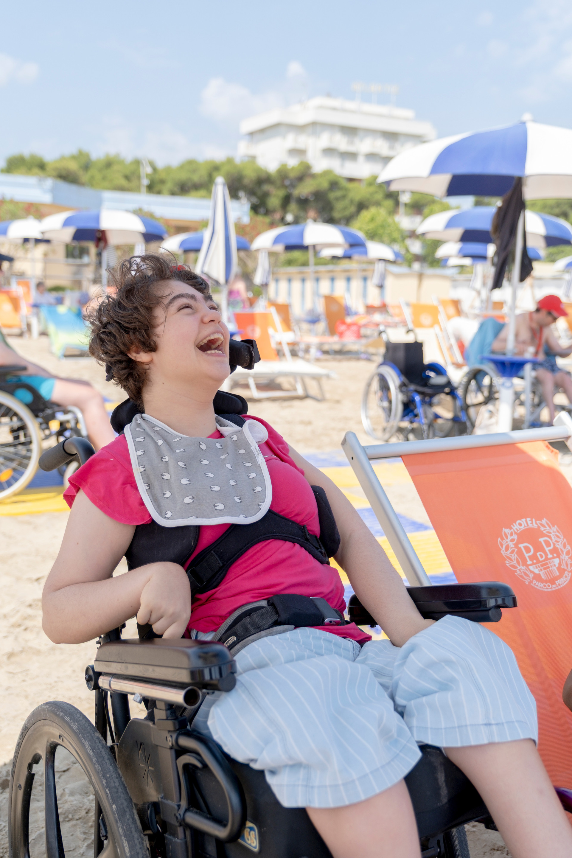 Una ragazza in carrozzina sorride felice in spiaggia durante il soggiorno estivo organizzato dalla Lega del Filo d'Oro, vivendo momenti di gioia e inclusione.