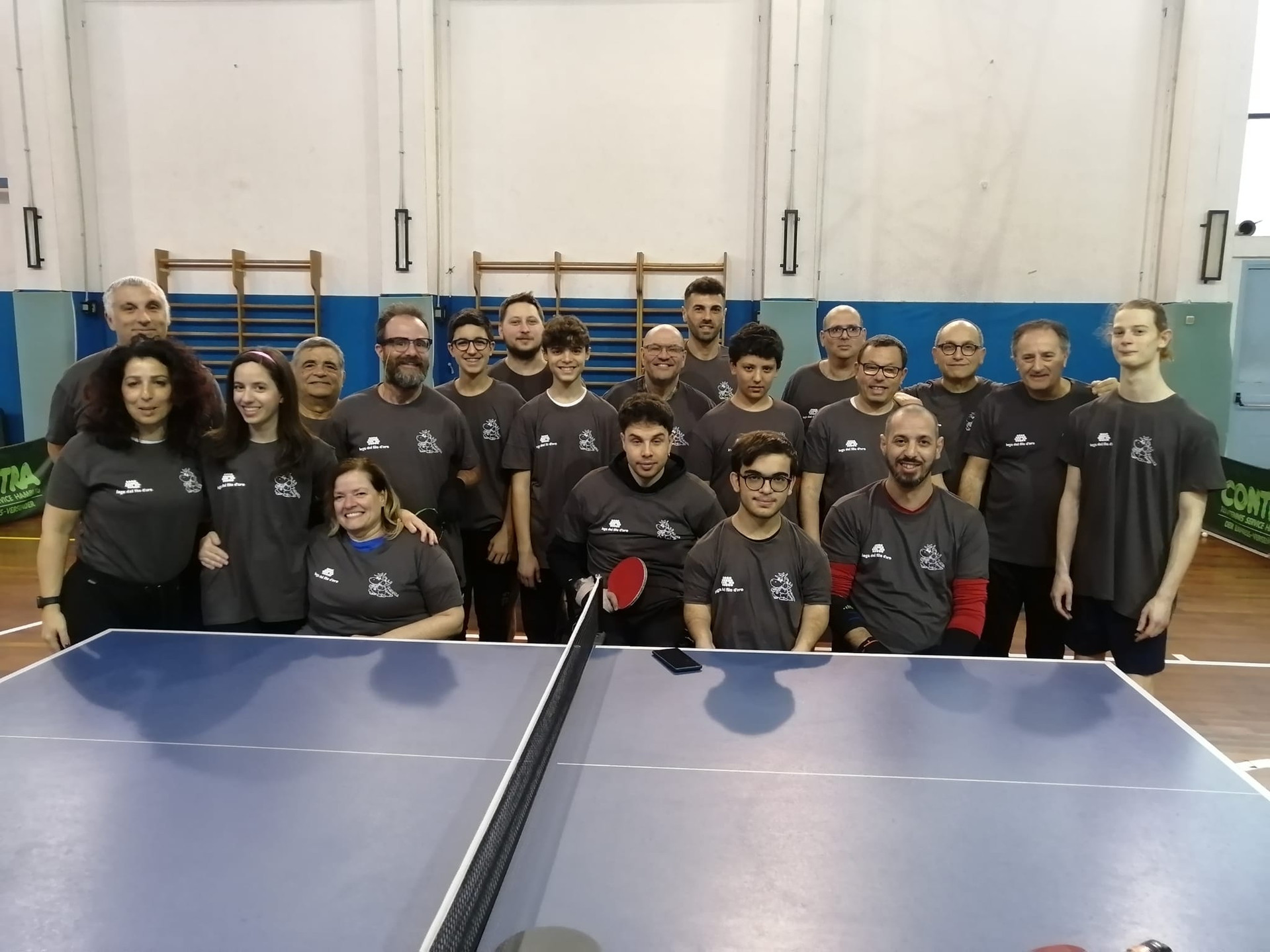 Gruppo di persone in palestra riunite dietro un tavolo da ping pong, tutte con la stessa maglietta grigia. Clima amichevole e sportivo