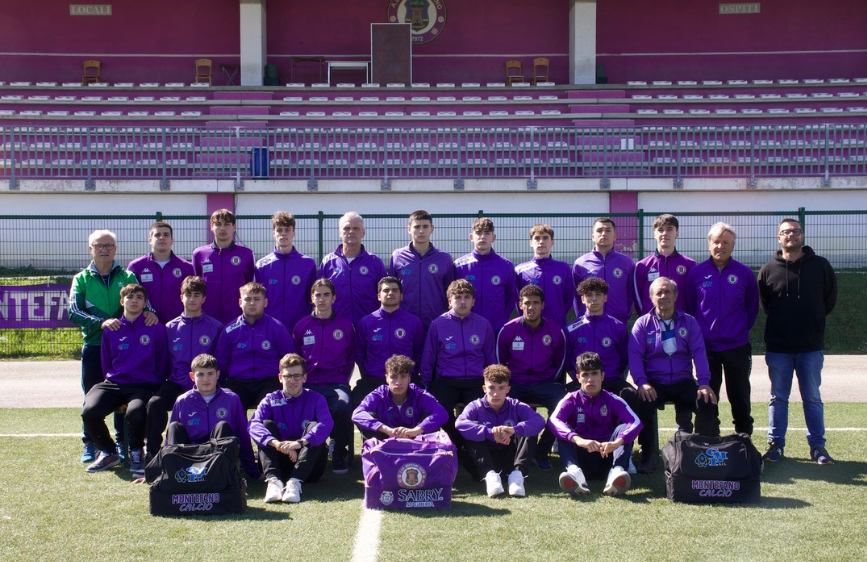  L'immagine mostra la squadra di calcio SSD Montefano Calcio: un gruppo di giovani calciatori e alcuni adulti posa per una foto di squadra su un campo da calcio. Indossano tutti delle divise viola e sono disposti su due file, con alcuni seduti in prima fila, di fronte a delle tribune viola.