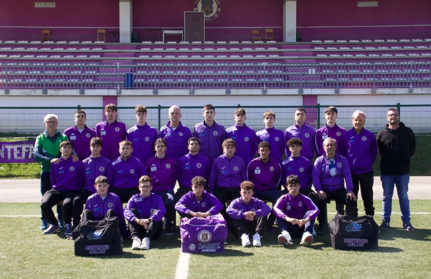  L'immagine mostra la squadra di calcio SSD Montefano Calcio: un gruppo di giovani calciatori e alcuni adulti posa per una foto di squadra su un campo da calcio. Indossano tutti delle divise viola e sono disposti su due file, con alcuni seduti in prima fila, di fronte a delle tribune viola.