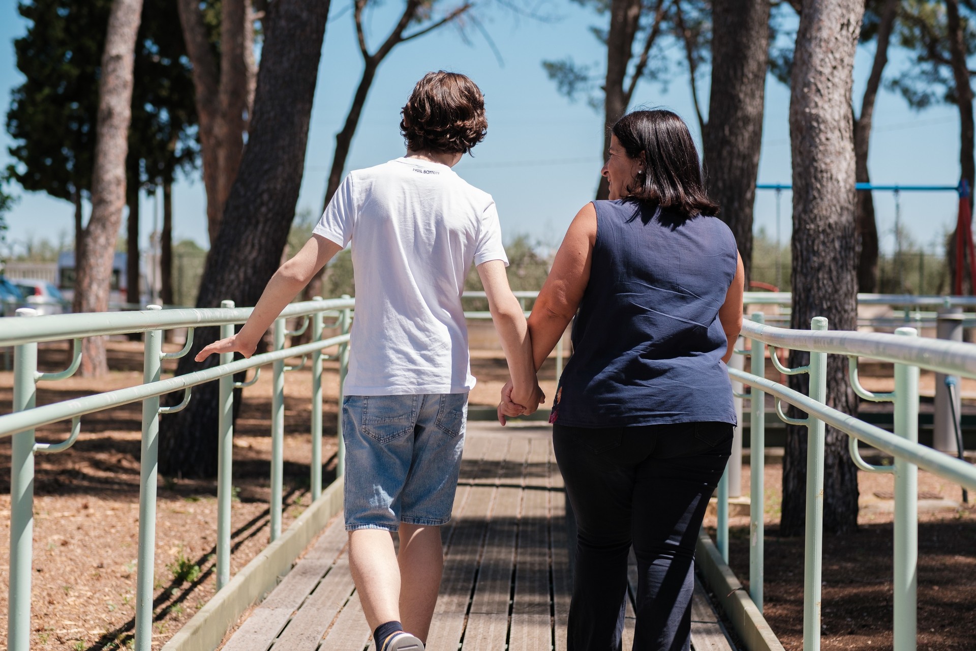 Un utente e una volontaria della Lega del Filo d'Oro camminano di spalle lungo una passerella di legno immersa nel verde di un parco all’aperto. Procedono affiancati, mano nella mano, circondati da alberi.