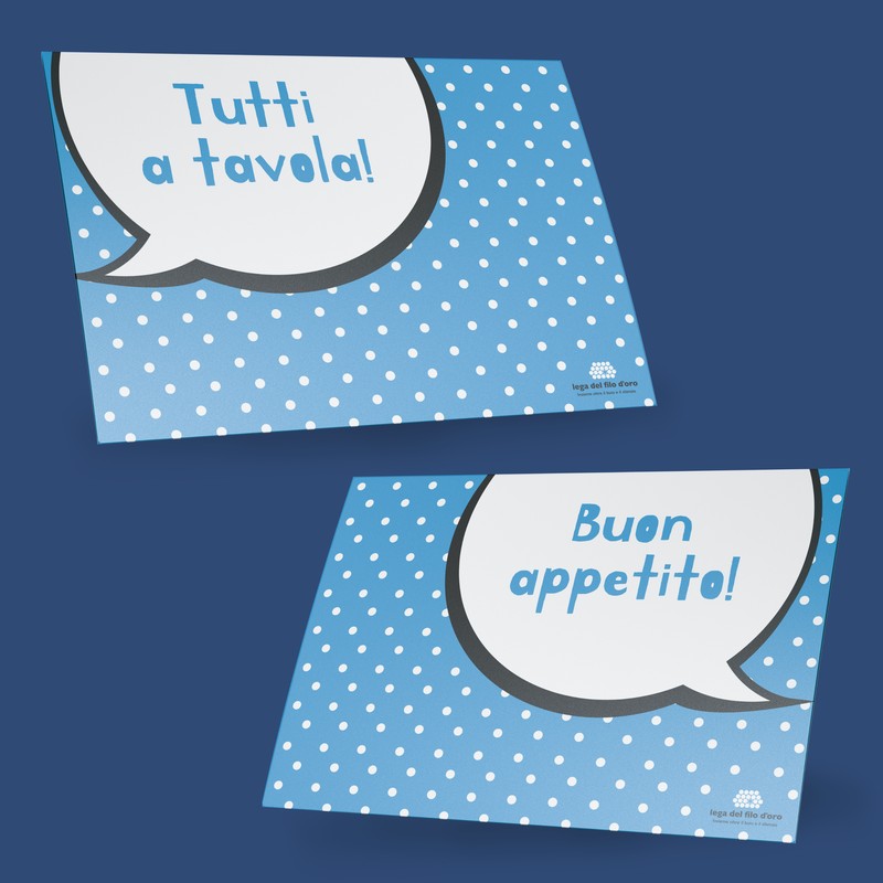 Due tovagliette da colazione in PVC azzurro con stampa a pois bianchi e la grafica di un fumetto - su una la frase è "Buon appetito!", sull'altra "Tutti a tavola!". Su entrambe in un angolo si trova il logo della Fondazione 