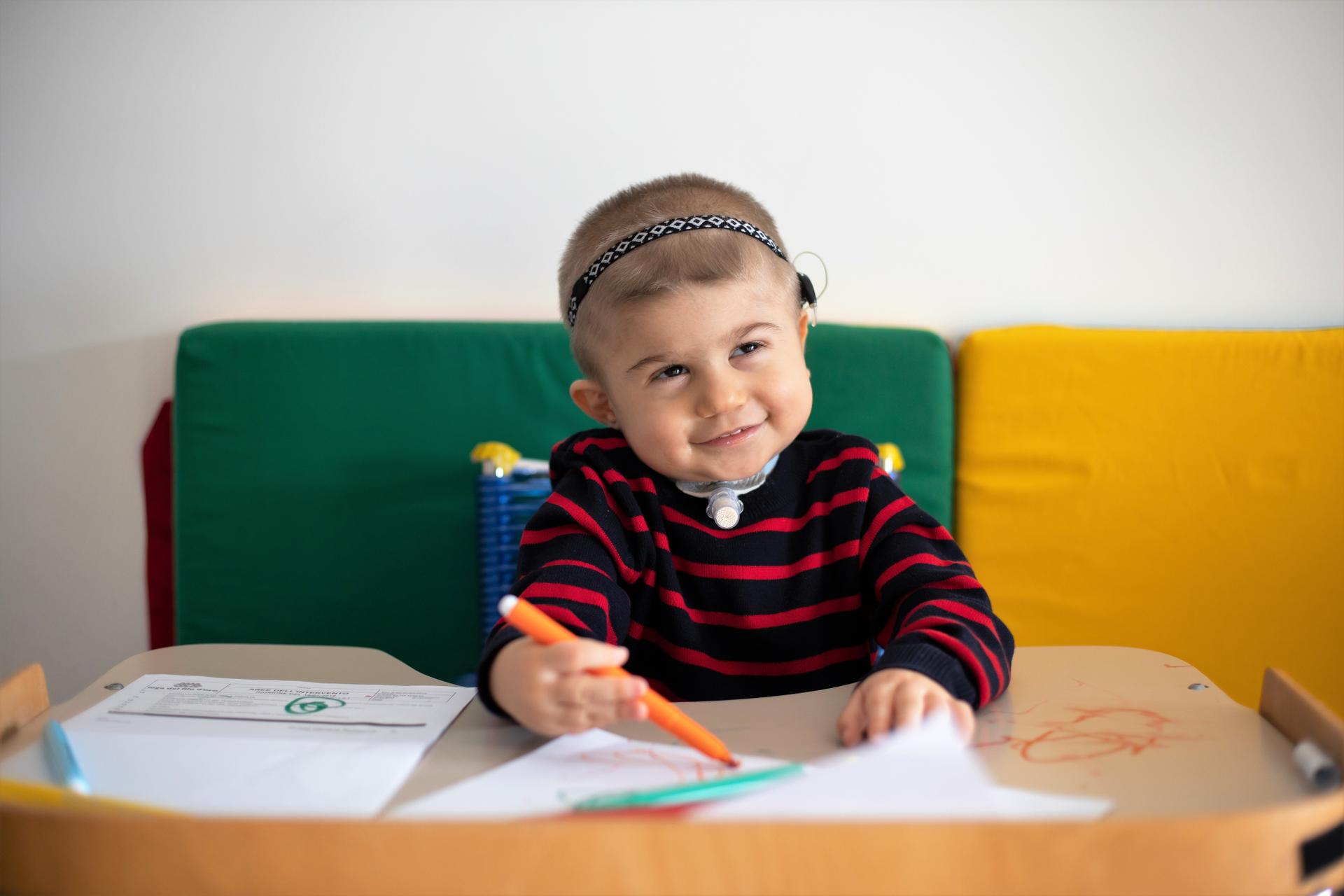 Berk, un bambino, utente della Lega del Filo d'Oro, sorride mentre impugna un pennarello arancione per fare un disegno.