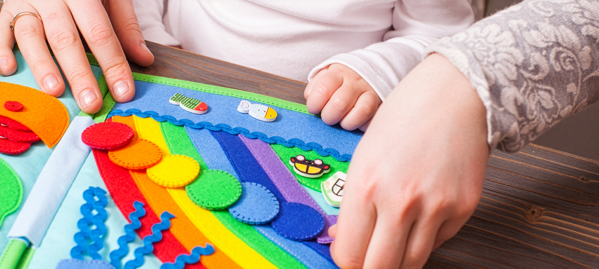 Nell'immagine compare un libro tattile raffigurante un arcobaleno realizzato con la stoffa colorata. Le mani di una donna adullta e di una bambina interagiscono con il libro. 
