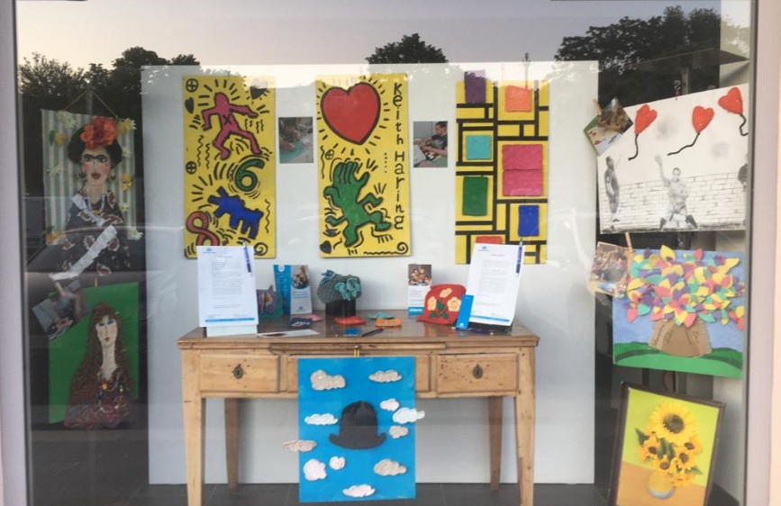 Vetrina con opere colorate ispirate a Keith Haring, Mondrian e Magritte, realizzate da utenti della Lega del Filo d’Oro. I quadri sono esposti attorno a un tavolo in legno chiaro con materiali informativi.