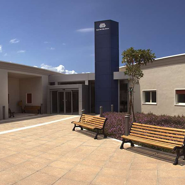 Grandangolo del Centro di Termini Imerese. L'ingresso piastrellato è centrale, due panchine e un piccolo giardino con dei fiori e due alberelli sono sulla destra, mentre l'edificio è grigio. Il cielo è azzurro.
