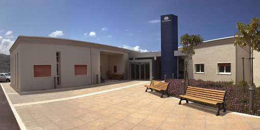 Grandangolo del Centro di Termini Imerese. L'ingresso piastrellato è centrale, due panchine e un piccolo giardino con dei fiori e due alberelli sono sulla destra, mentre l'edificio è grigio. Il cielo è azzurro.