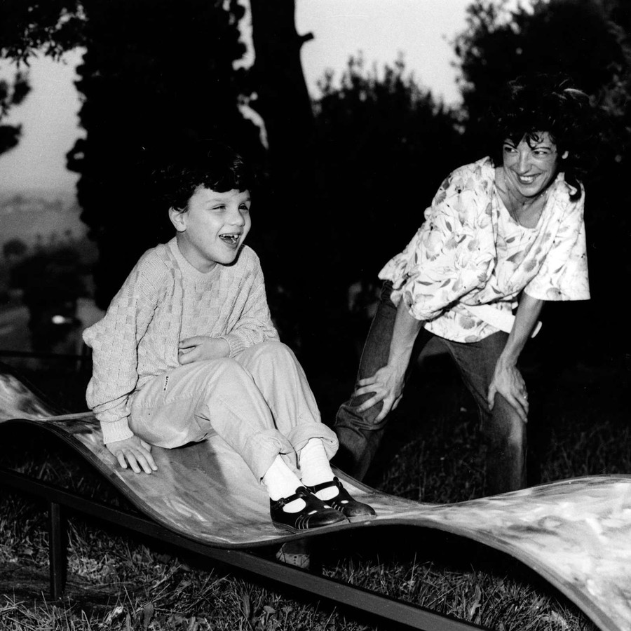 Un bambino gioca e ride su uno scivolo all'aperto, mentre un donna lo guarda e ride. Immagine in bianco e nero.