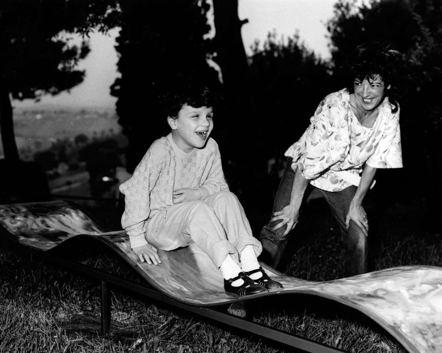 Un bambino gioca e ride su uno scivolo all'aperto, mentre un donna lo guarda e ride. Immagine in bianco e nero.