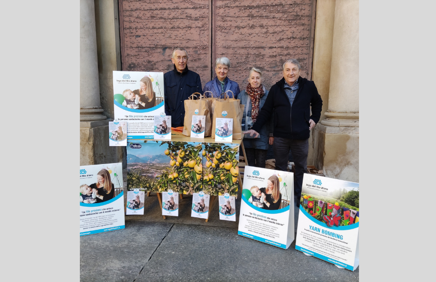L'immagine mostra un gruppo di quattro persone anziane, sorridenti, in piedi dietro un tavolo allestito con materiale promozionale. Sul tavolo ci sono piante di agrumi con frutti gialli, borse di carta marrone e diversi volantini o poster che mostrano una donna con un bambino e riferimenti a "Yarn Bombing".