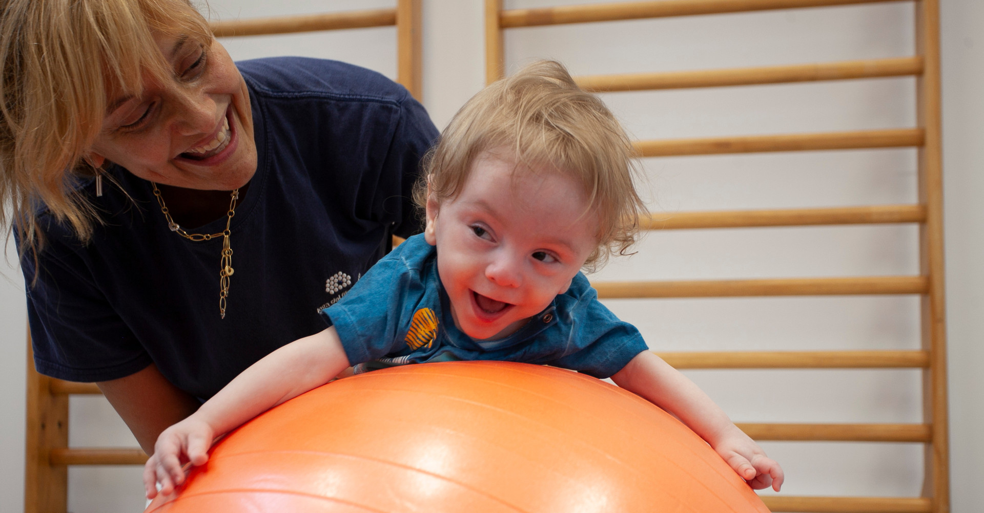 Gabriele, un bambino seguito dalla Lega del Filo d'Oro fa attività di fisioterapia in palestra con la sua fisioterapista, che lo sorregge in equilibrio sdraiato su una grande palla arancione. Entrambi ridono divertiti.