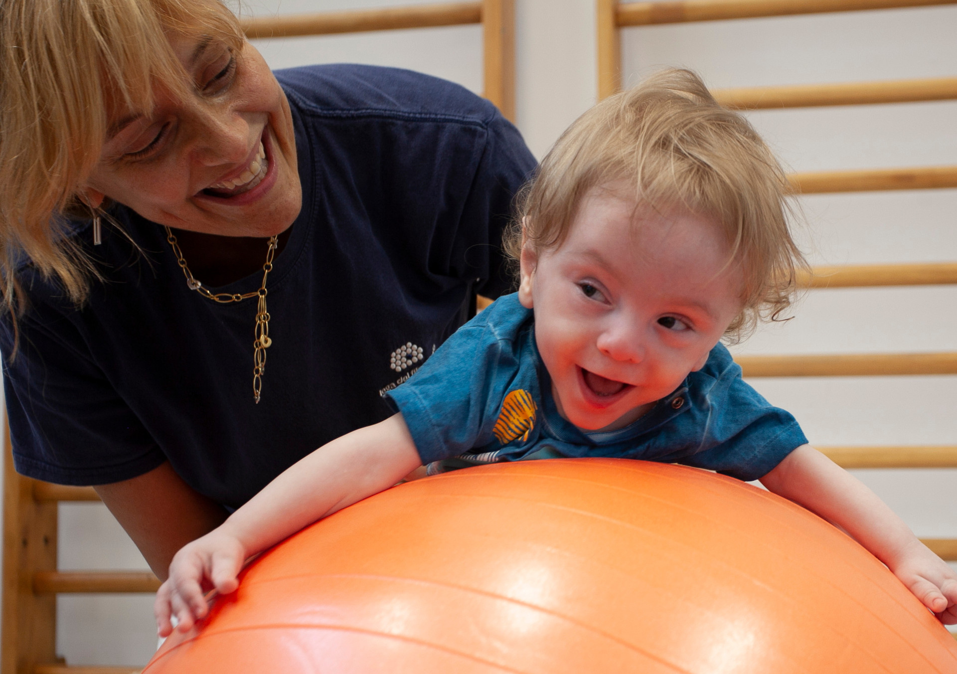 Gabriele, un bambino seguito dalla Lega del Filo d'Oro fa attività di fisioterapia in palestra con la sua fisioterapista, che lo sorregge in equilibrio sdraiato su una grande palla arancione. Entrambi ridono divertiti.