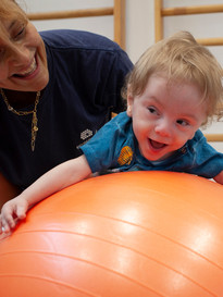 Gabriele, un bambino seguito dalla Lega del Filo d'Oro fa attività di fisioterapia in palestra con la sua fisioterapista, che lo sorregge in equilibrio sdraiato su una grande palla arancione. Entrambi ridono divertiti.