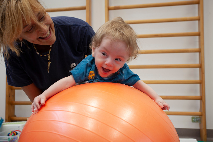 Gabriele, un bambino seguito dalla Lega del Filo d'Oro fa attività di fisioterapia con la sua fisioterapista, che lo sorregge in equilibrio sdraiato su una grande palla arancione. Entrambi ridono divertiti. 