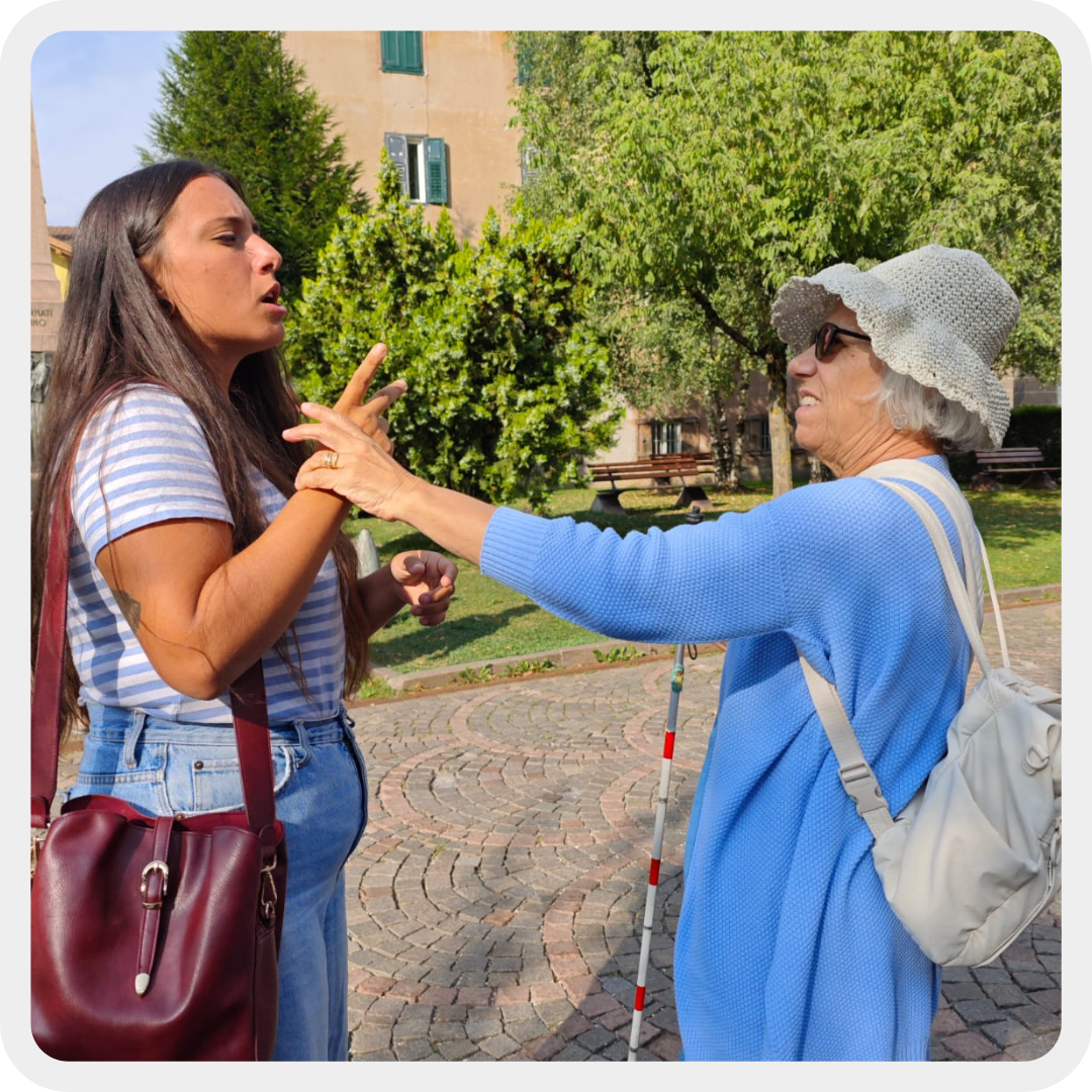 Nella foto, un utente sordocieca tiene con una mano il bastone bianco e rosso e con l'altra si appoggia alla mano della volontaria che le sta parlando tramite LIS tattile