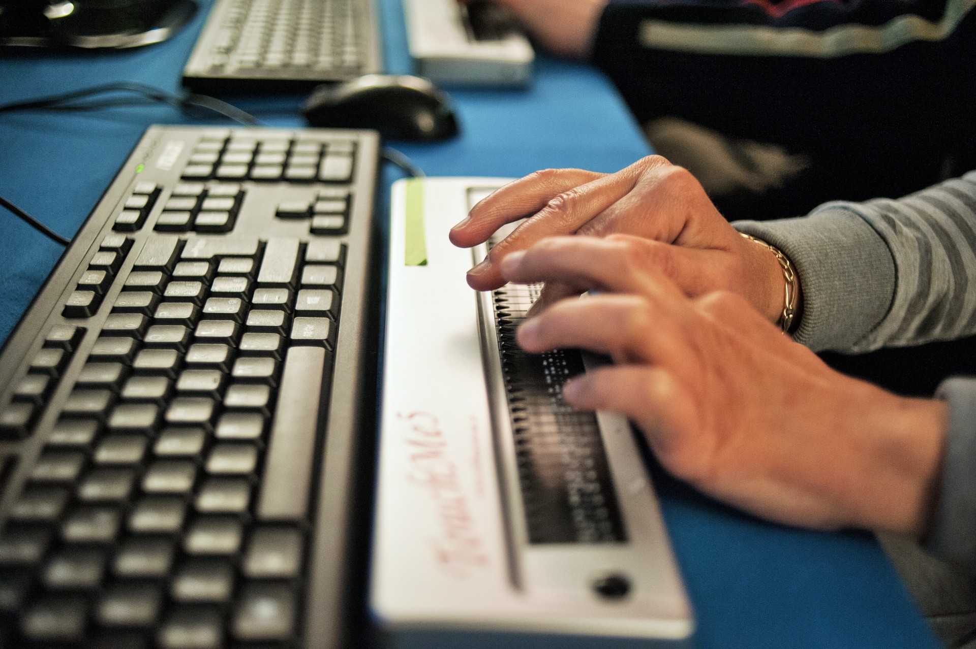 Dettaglio di due mani che scorrono su una barra braille appoggiata su una scrivania. Davanti alla barra è posizionata la tastiera di un computer.