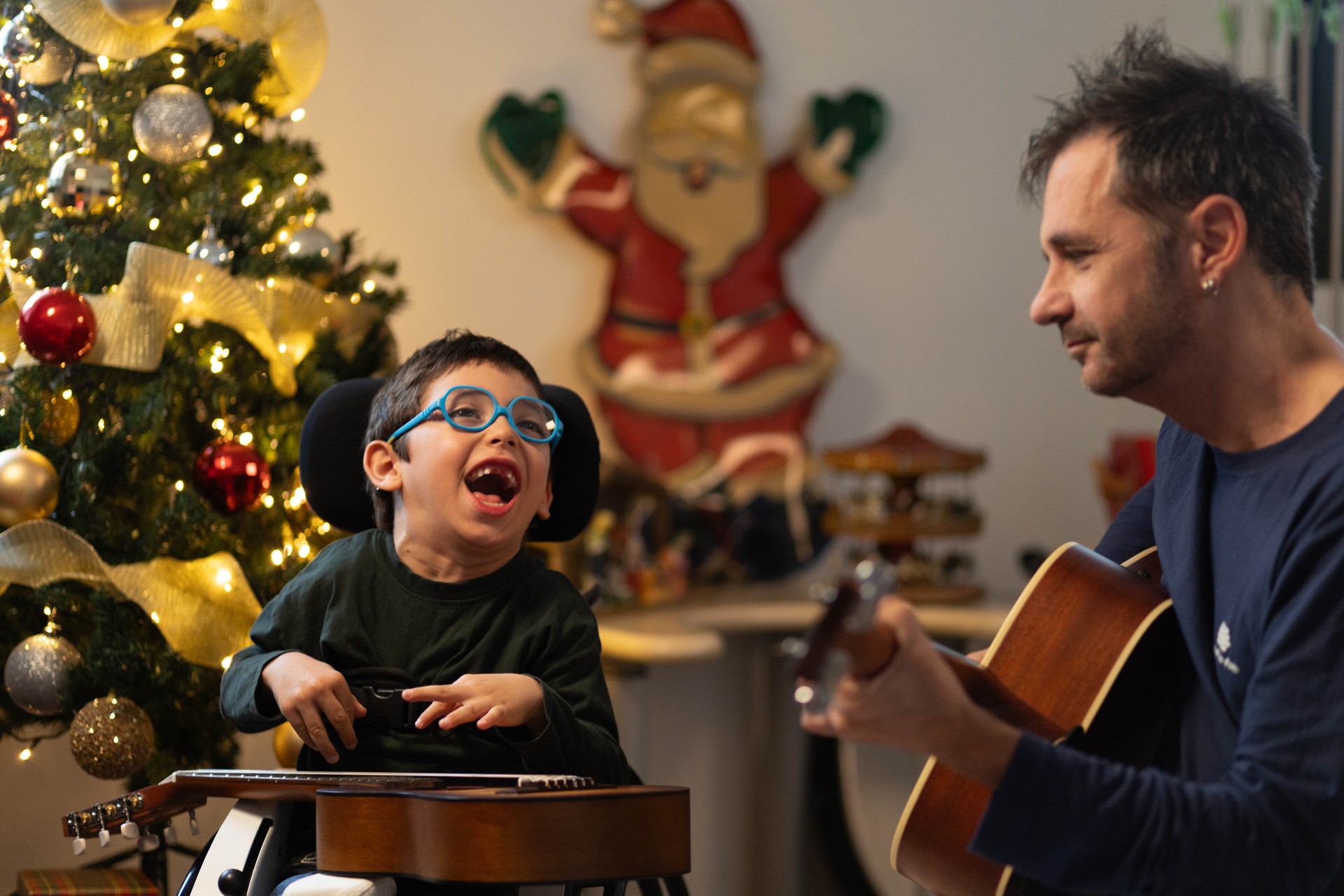Nicola, un bimbo con ipovisione e difficoltà motorie, insieme al musicoterapista che suona la chiatarra. Sullo sfondo un albero di Natale addobbato. Nicola ride, è felice