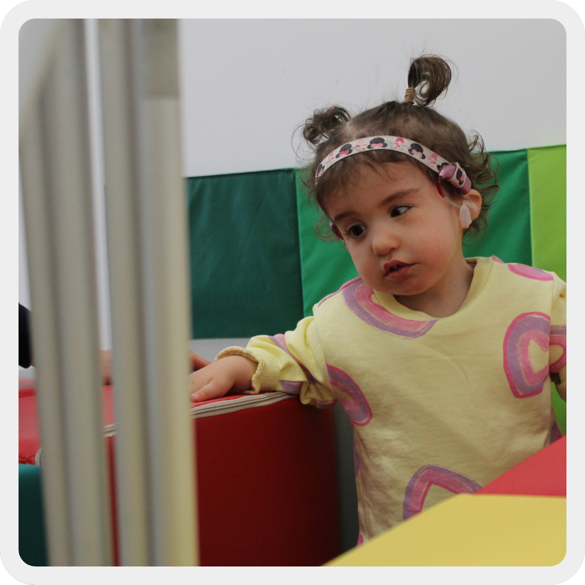 Sophie. bambina con Sindrome di Charge di circa 3 anni, si sorregge a cubi di gomma colorati mentre muove qualche passo. Indossa una felpa gialla e rosa, ha l'impianto cocleare. 